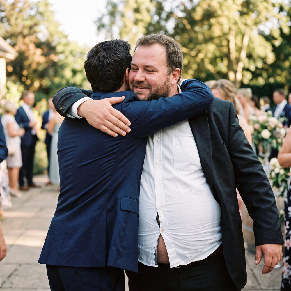 Big man at a wedding hugs another guest while his white dress shirt gaps open across the stomach, exposing his belly as the buttons pull apart during the embrace. The candid moment highlights a common button-gap problem in regular dress shirts for larger men.